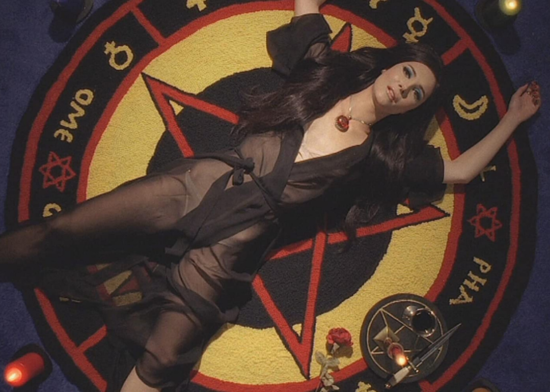 A dark haired woman in a transparent robe lays on a circular rug covered in pagan symbols with a knife next to her.