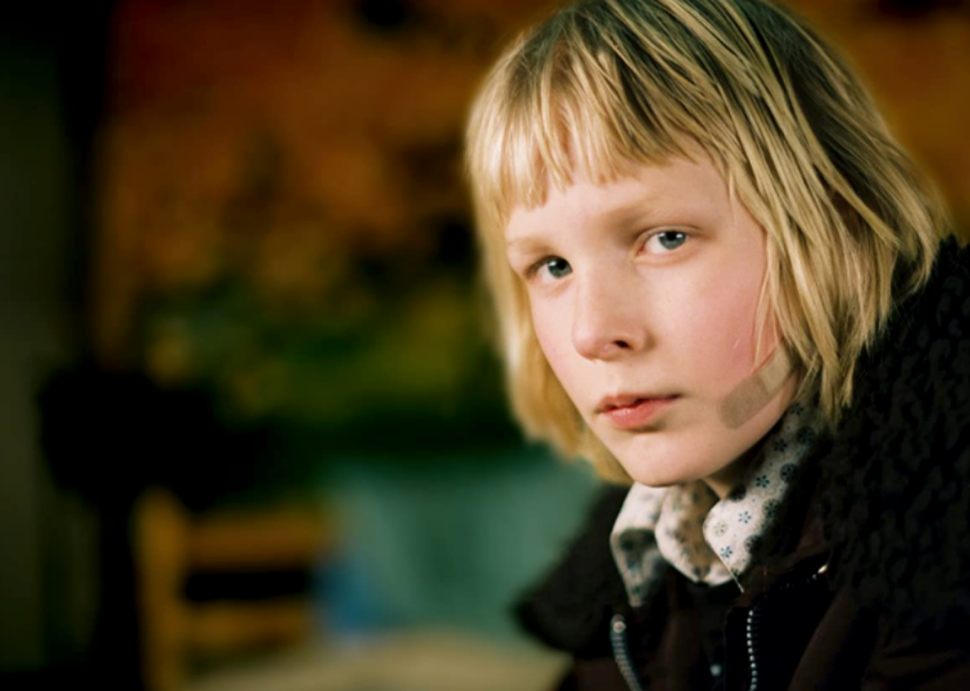 A young girl with short blonde hair and a bandaid on her face sits with a serious look.