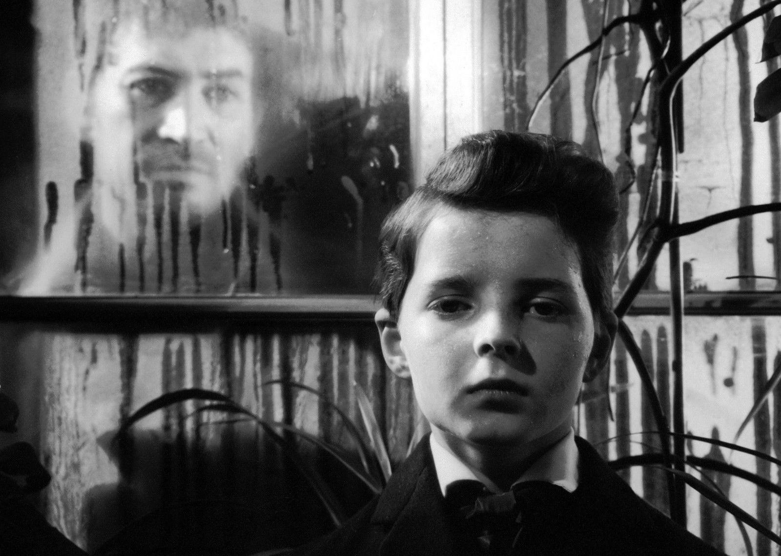 A ghostly man looking through a window at a young boy in a coat and tie.