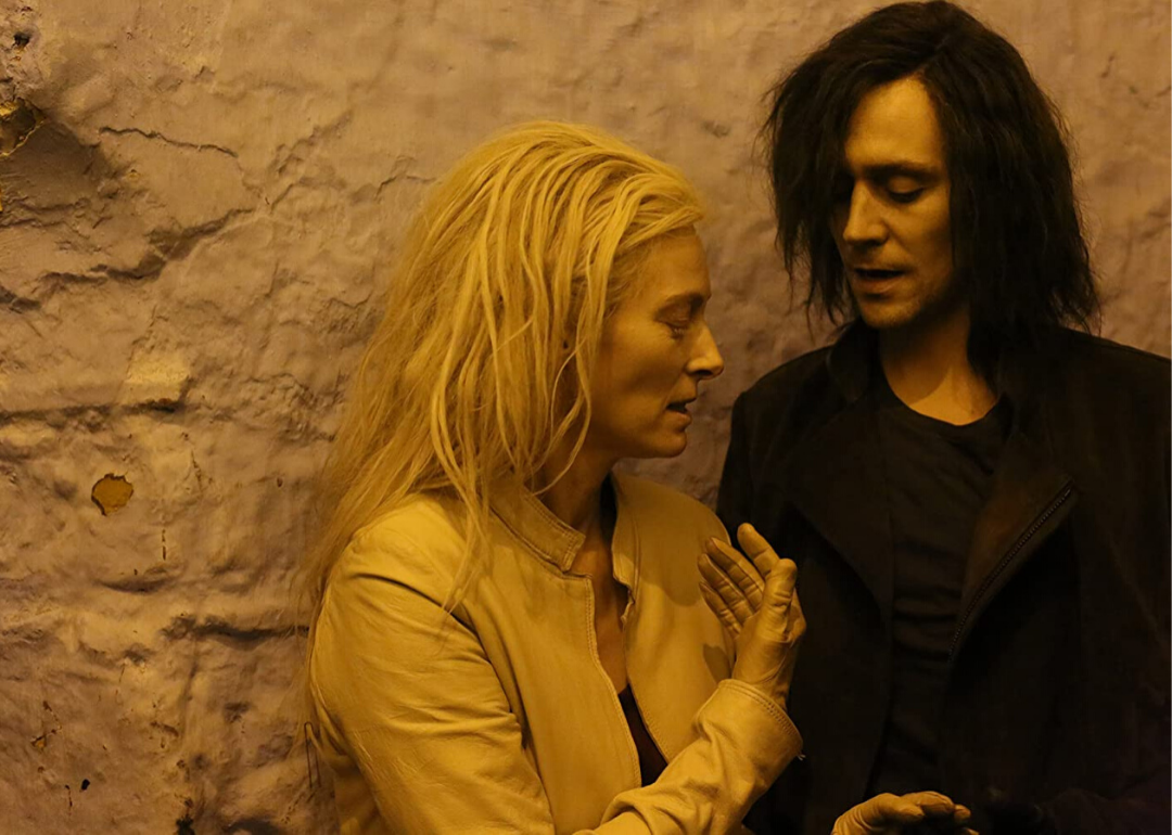 Tilda Swinton and Tom Hiddleston stand together talking, her with a leather jacket and gloves on and he wearing all black with black long hair.