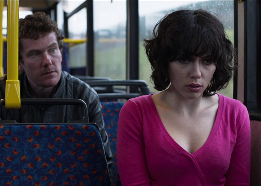 Scarlett Johansson in a black short wig sitting on a bus as a man sitting behind her watches her.