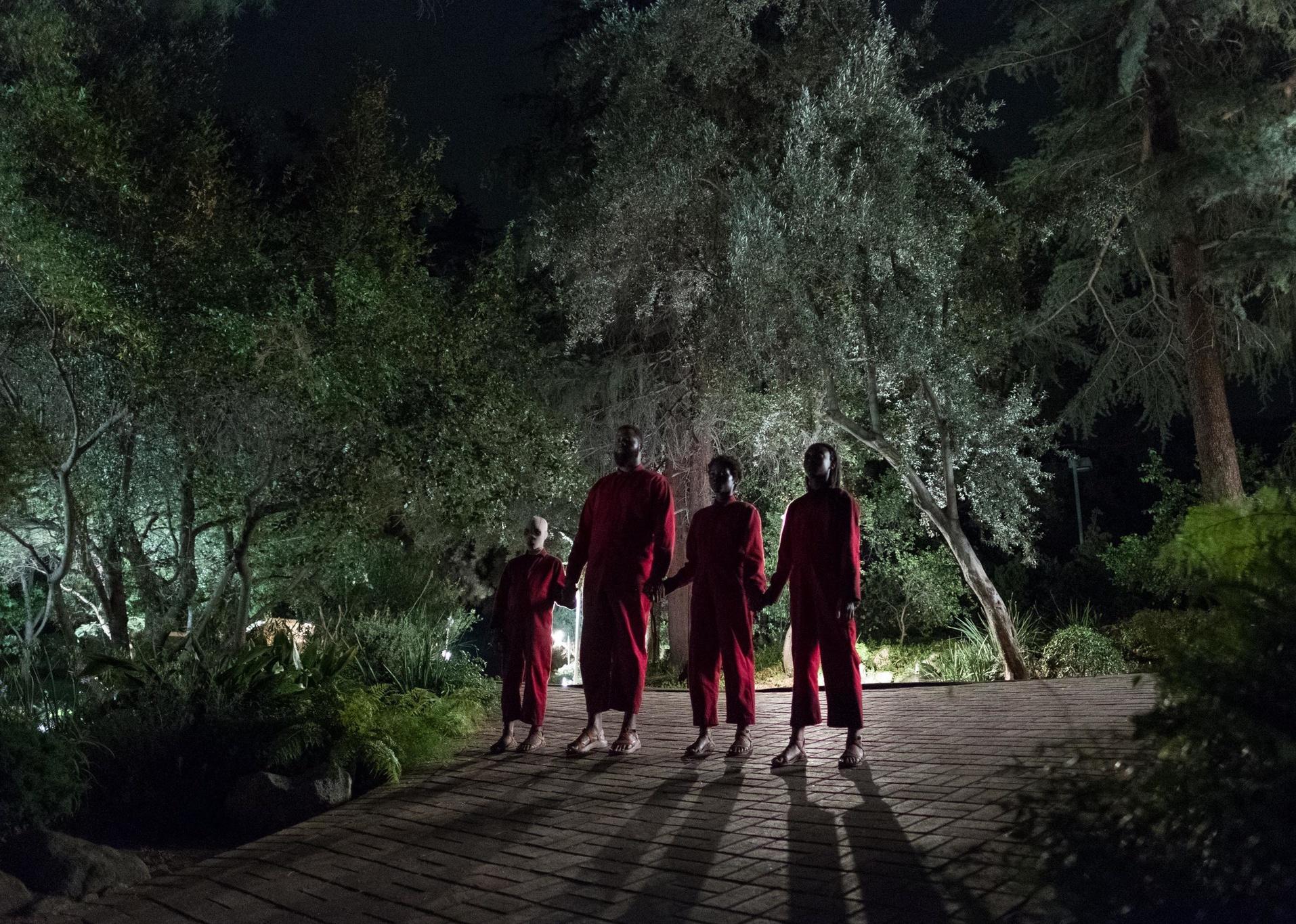 An older man and three young people stand holding hands wearing red jumpsuits in the dark.