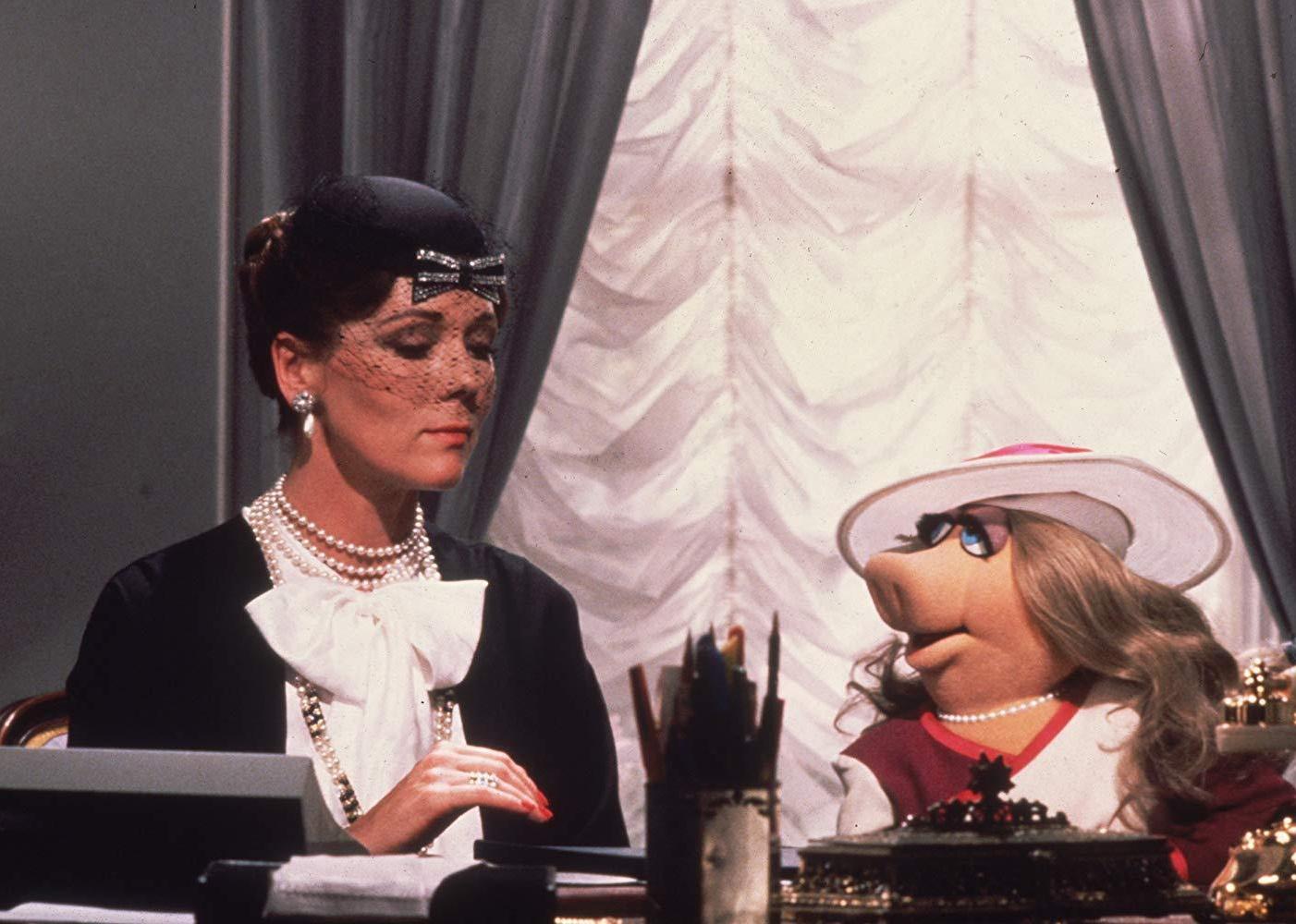 A woman in black and white dress sits at a desk with Miss Piggy the muppet.