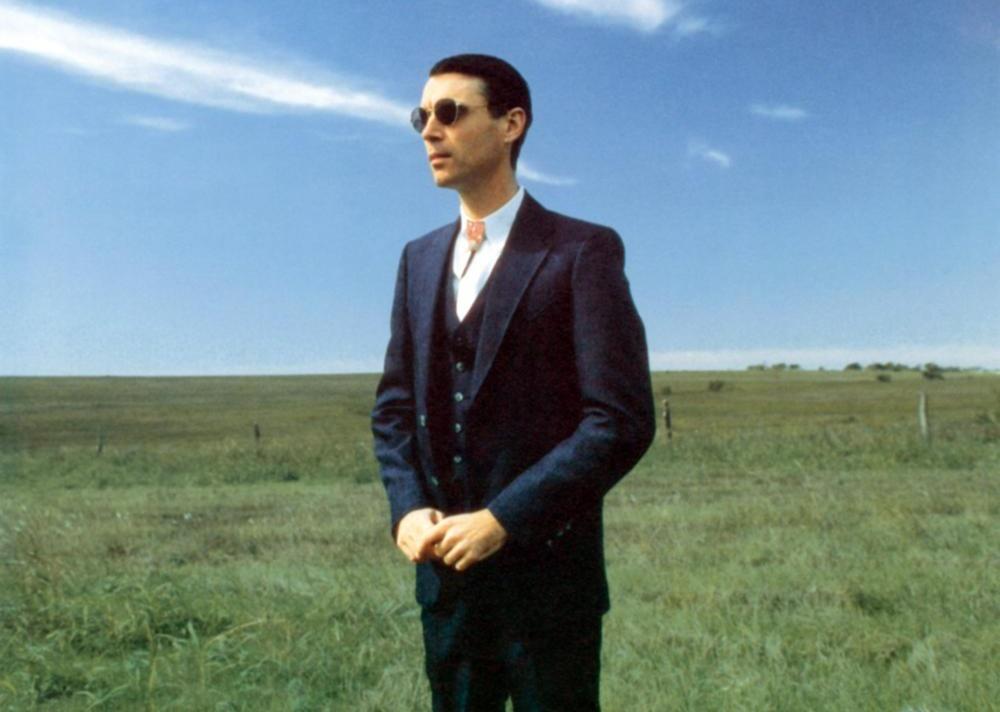David Byrne in a black suit standing in a field.