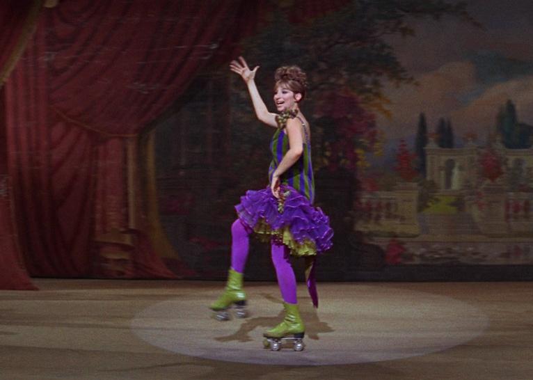 Barbra Streisand in a striped poofy dress rollerskating across a stage.