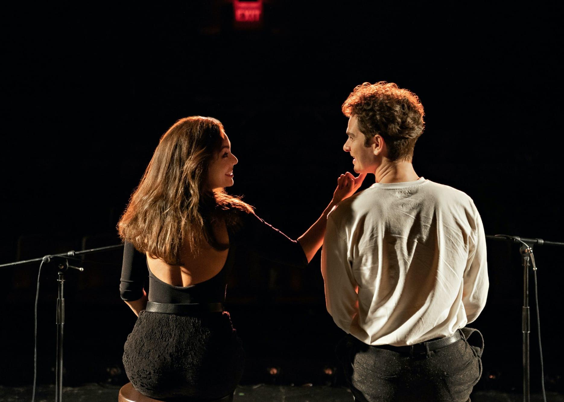 Vanessa Hudgens and Andrew Garfield sitting onstage towards a dark theater facing each other.