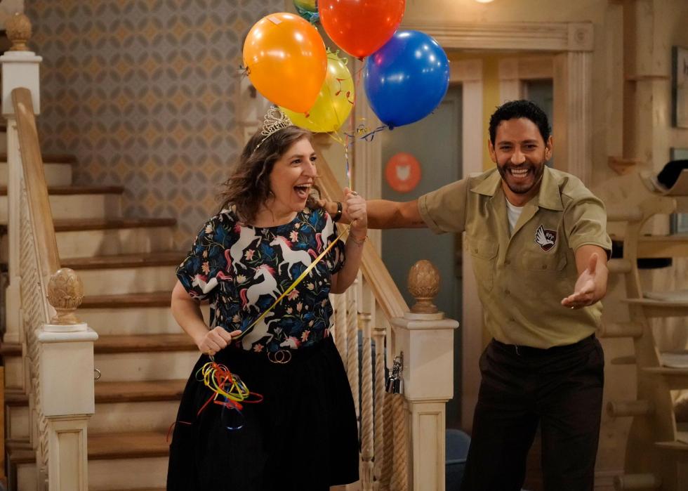 A woman looking surprised holds colorful balloons next to a man in a brown delivery uniform who is showing her something.