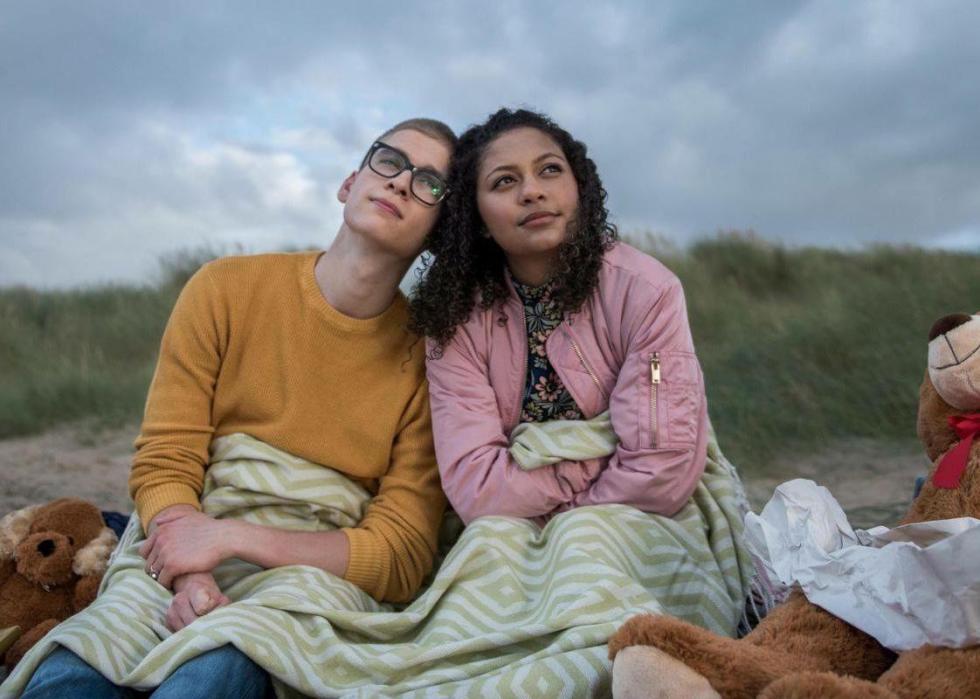 Two young people sit huddled together under a blanket on the beach.