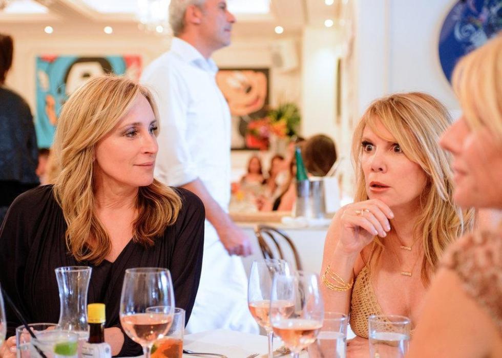 Women sit having wine at a restaurant, one looking shocked.
