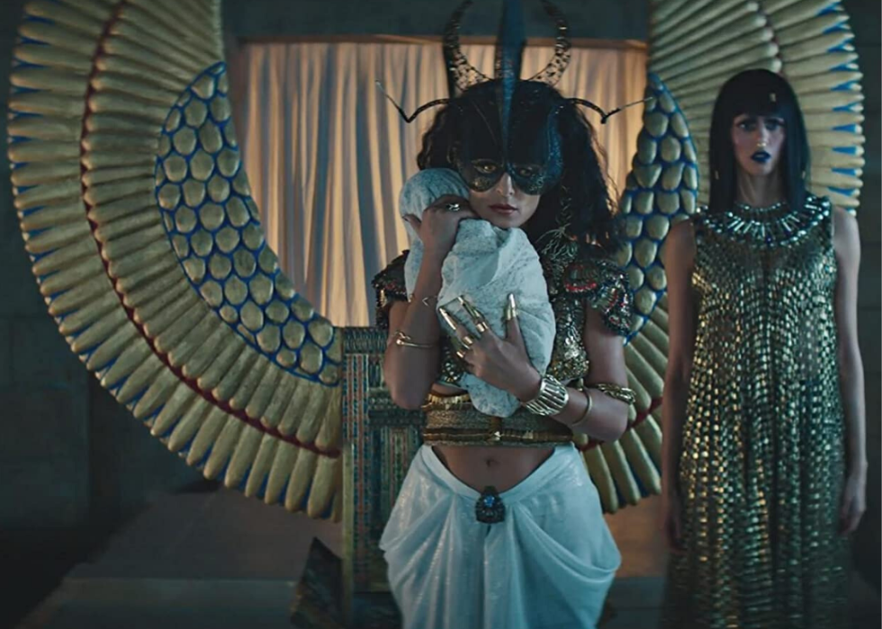 A woman in a mask holds a baby standing in front of large golden wings, next to another woman.