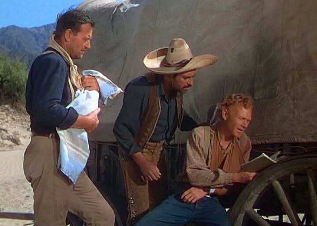 John Wayne, holding a baby, standing next to two cowboys next to a covered wagon.