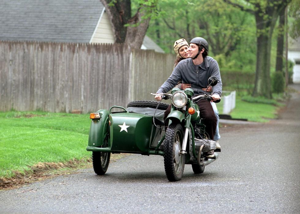A young man and woman ride on a green motorcycle with a sidecar.