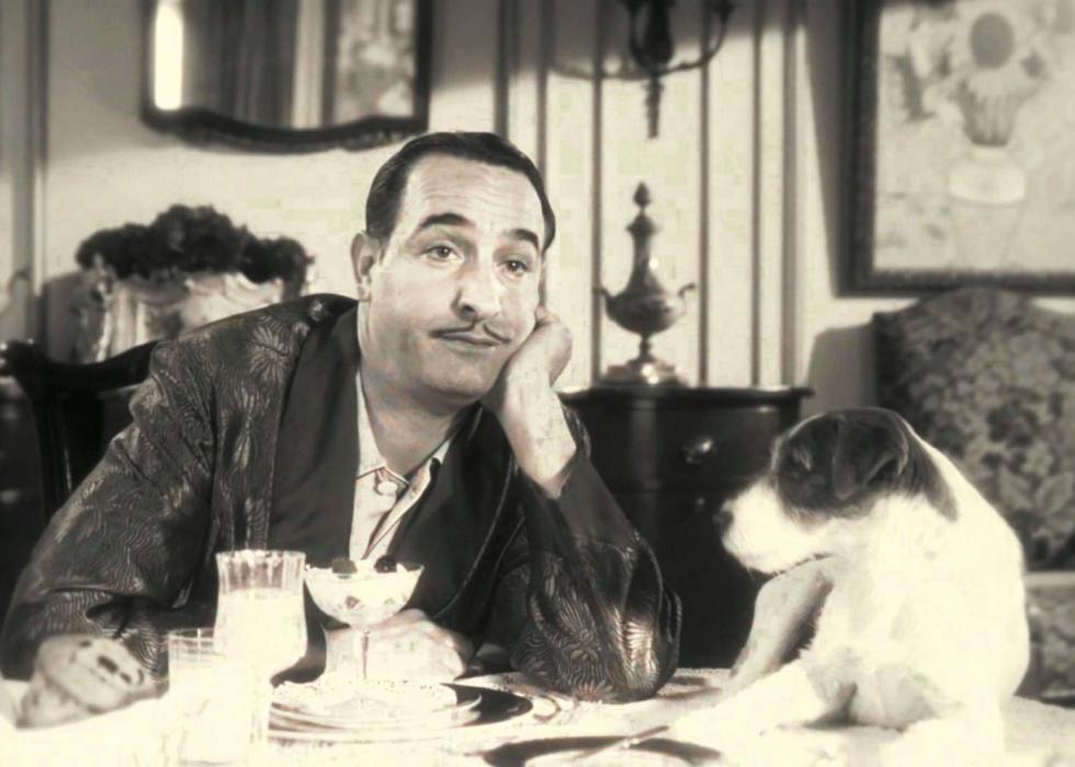 A man at a dinner table looking uninterested in his dessert with a dog next to him.