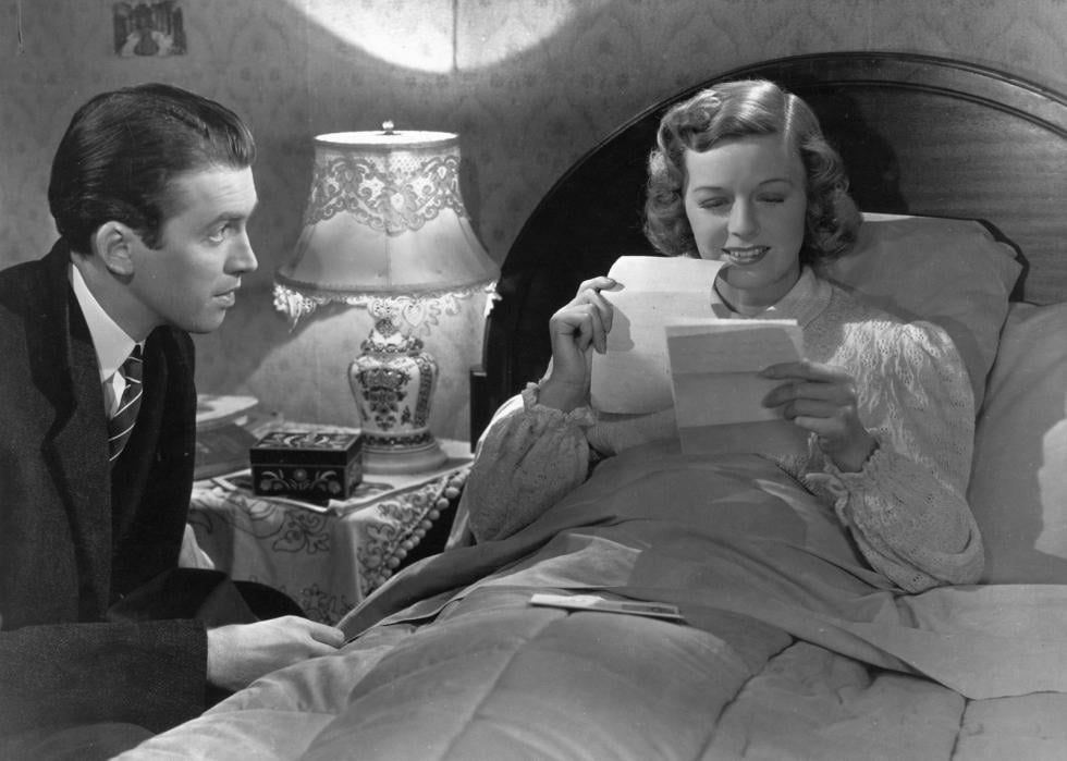 A woman lies in bed reading a letter and smiling while a man sits at her bedside.