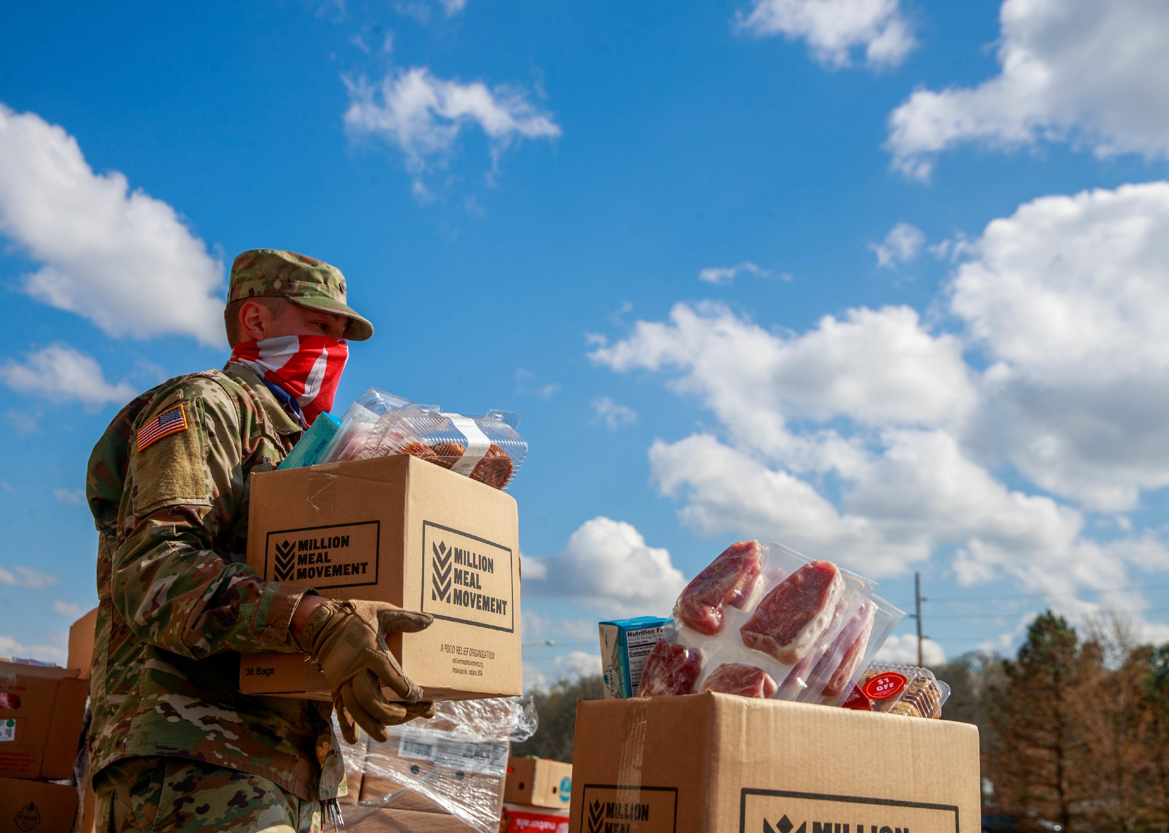 A member of the Indiana National Guard wearing an American flag balaclava carries a box of food while helping workers in distributing food.
