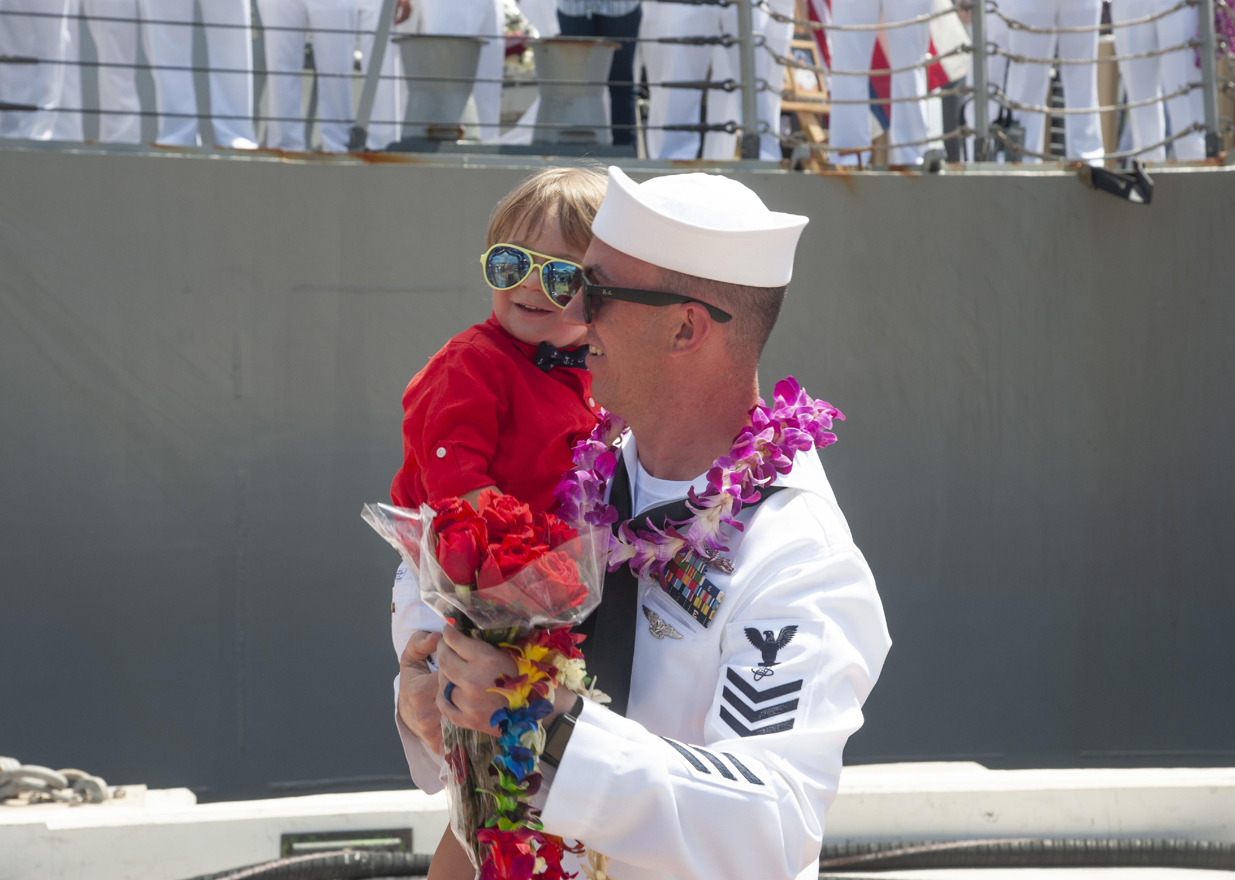 A sailor with flowers holds a little girl in sunglasses next to a ship.