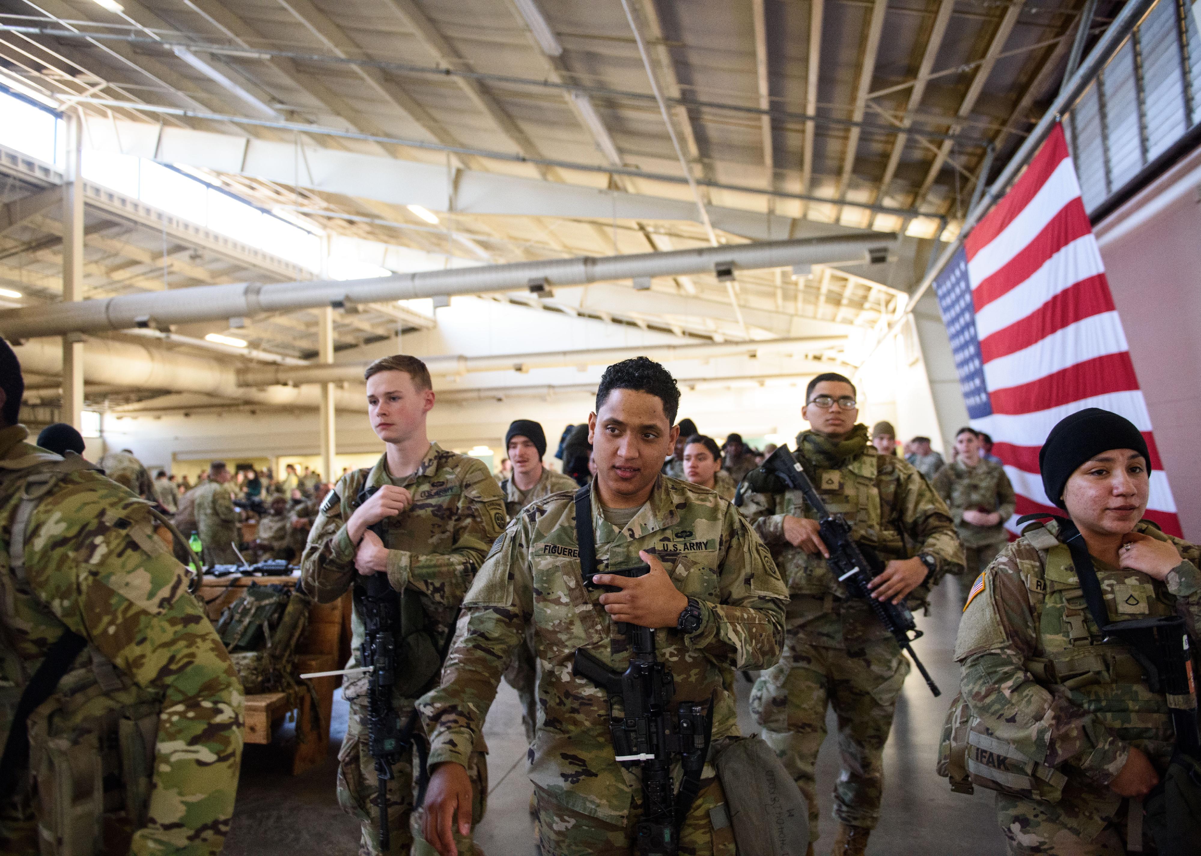 Soldiers in a warehouse listening to instruction next to a large American flag.