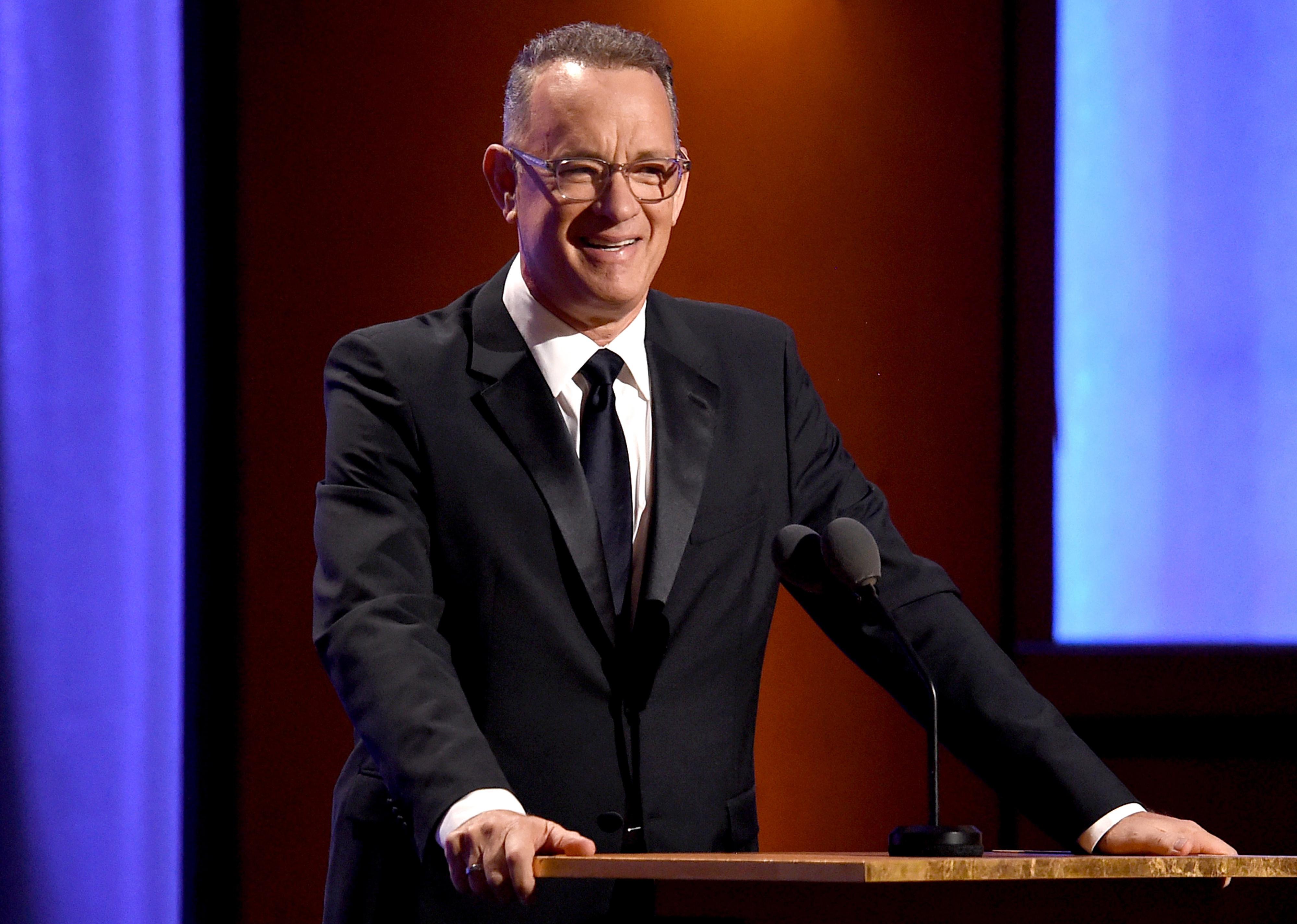 Tom Hanks in a black suit and tie onstage at a podium.