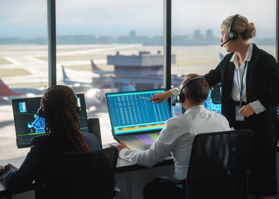 Air traffic controllers looking at computer screens in a tower overlooking a runway.