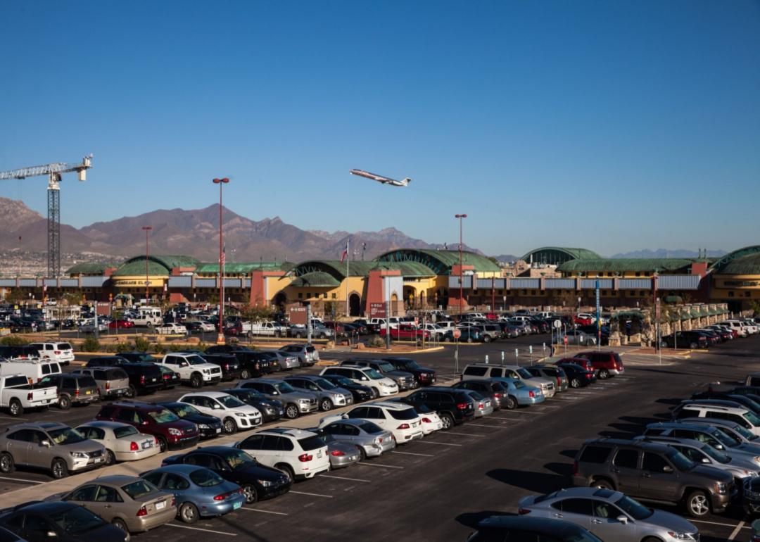 An airplane taking off over mountains behind the car-filled parking lot of El Paso airport.