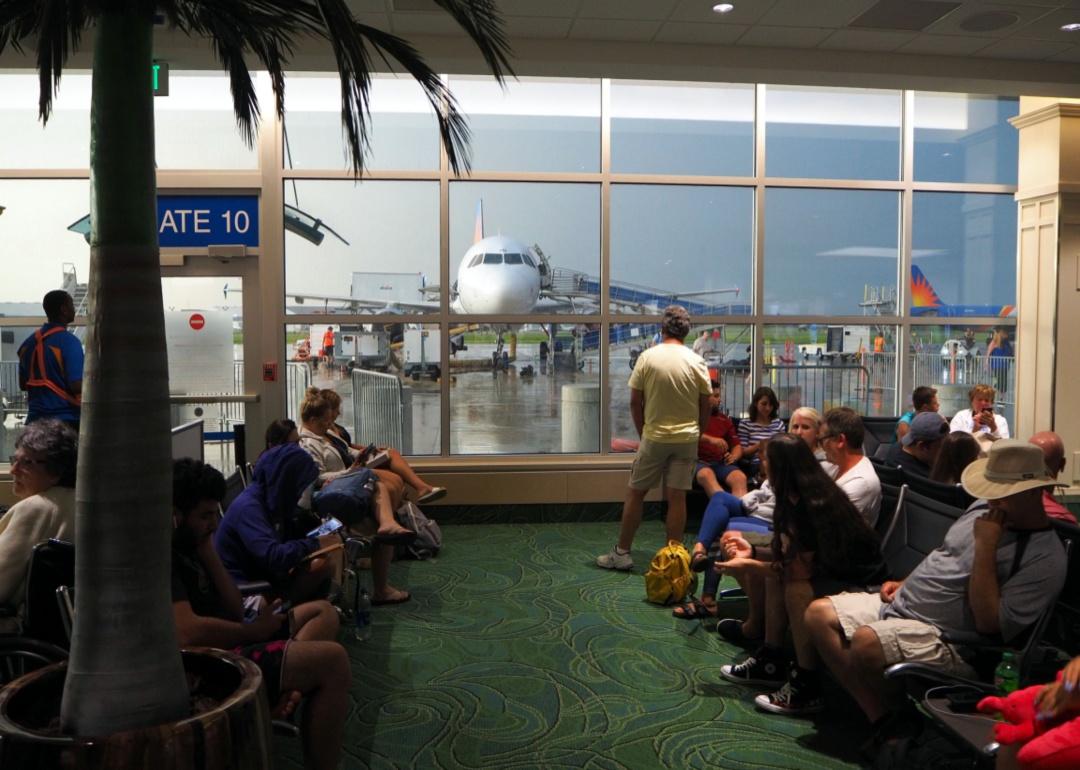 Passengers wait in a crowded room with a big window and palm tree facing a plane and storm outside on the tarmac.