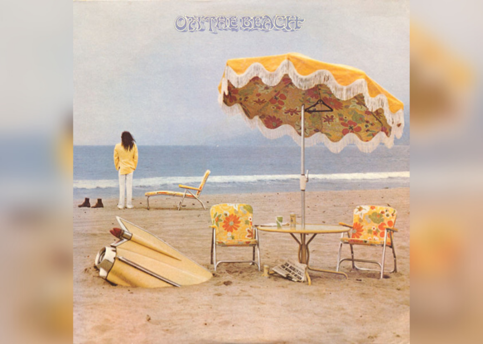 Neil Young standing on the beach in a yellow blazer looking at the ocean and everything on the beach is also yellow, including a table and chairs with umbrella..
