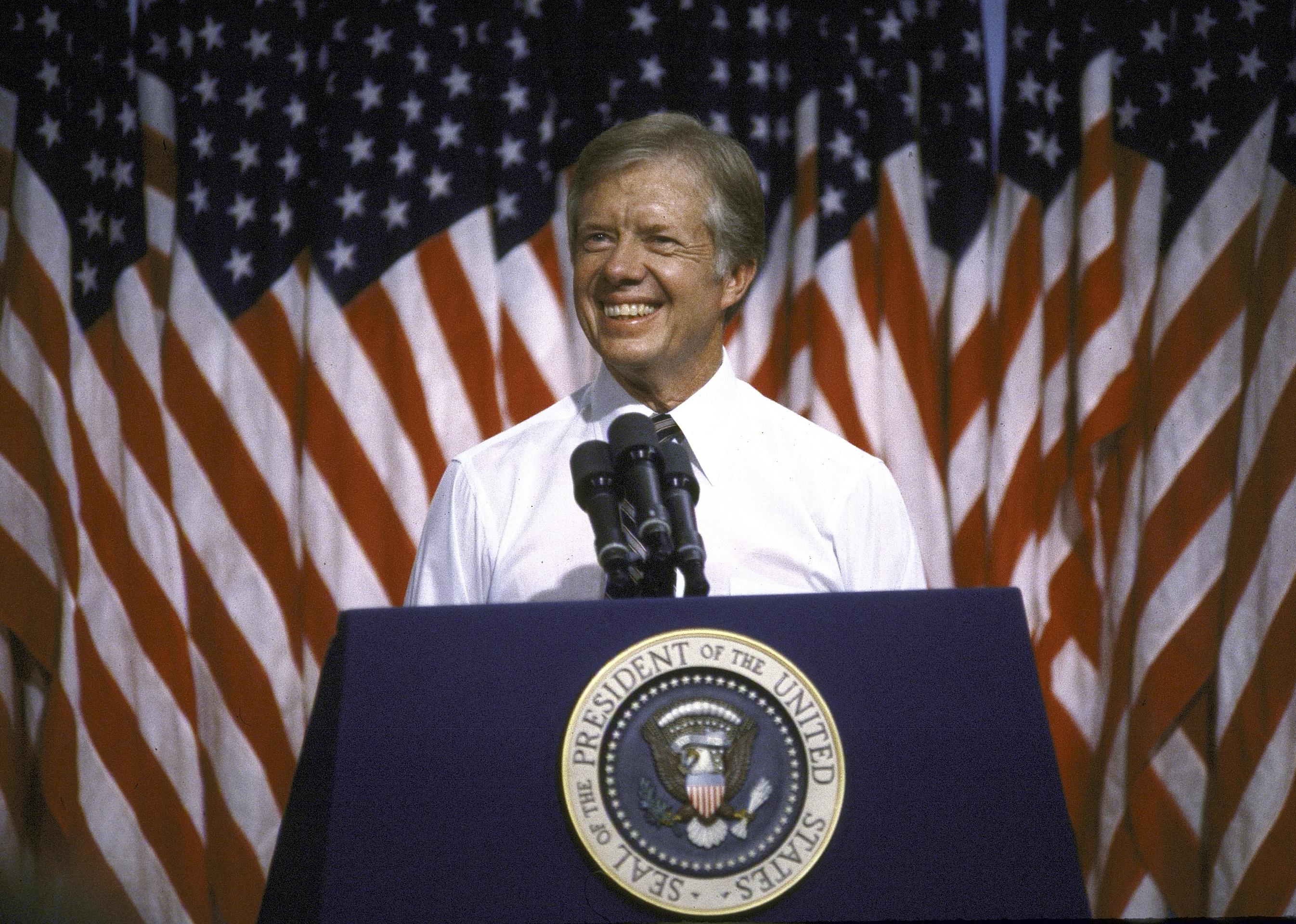 Jimmy Carter speaking at a presidential podium in front of American flags.