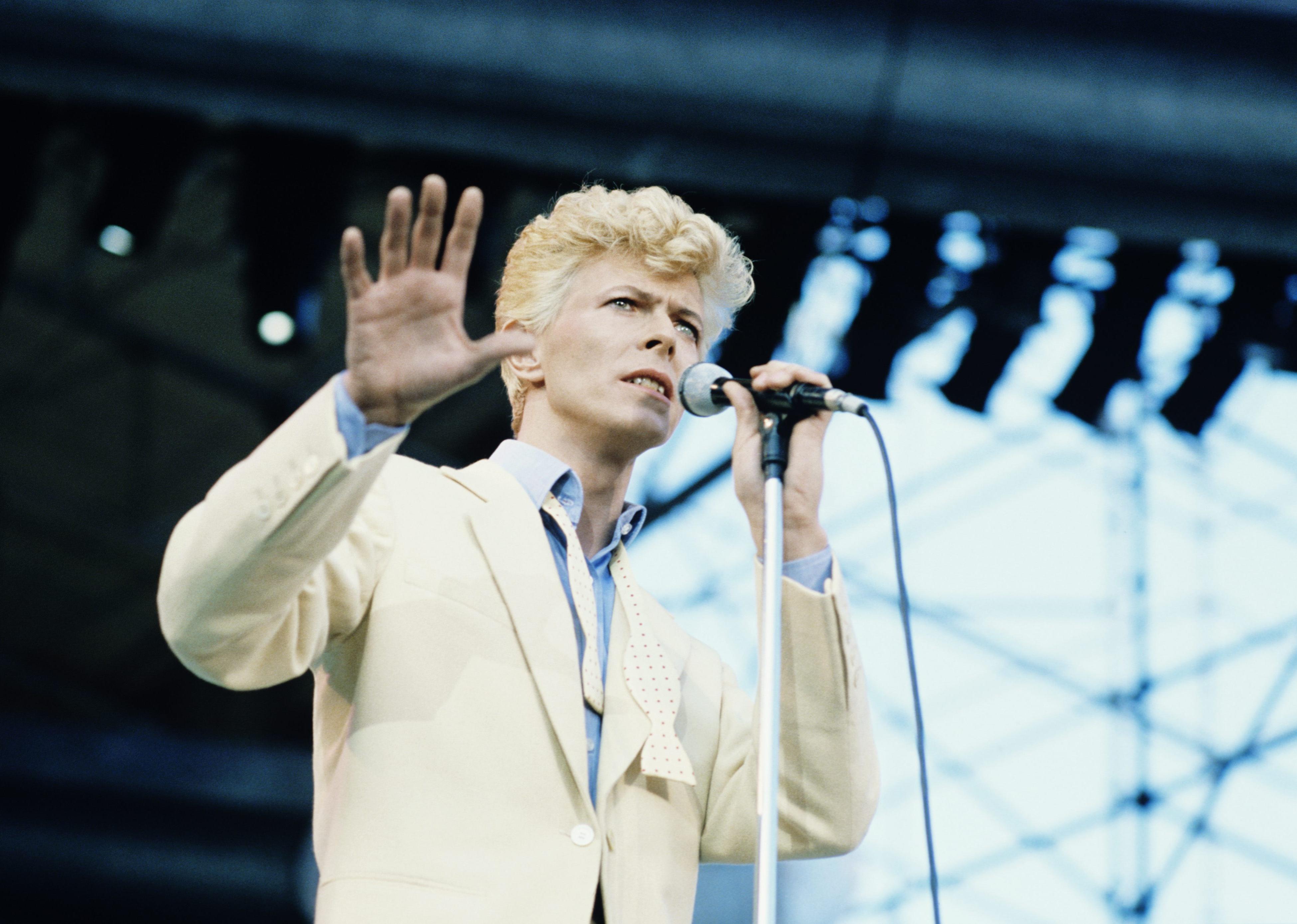 Bowie onstage in a cream colored suit jacket and undone tie.