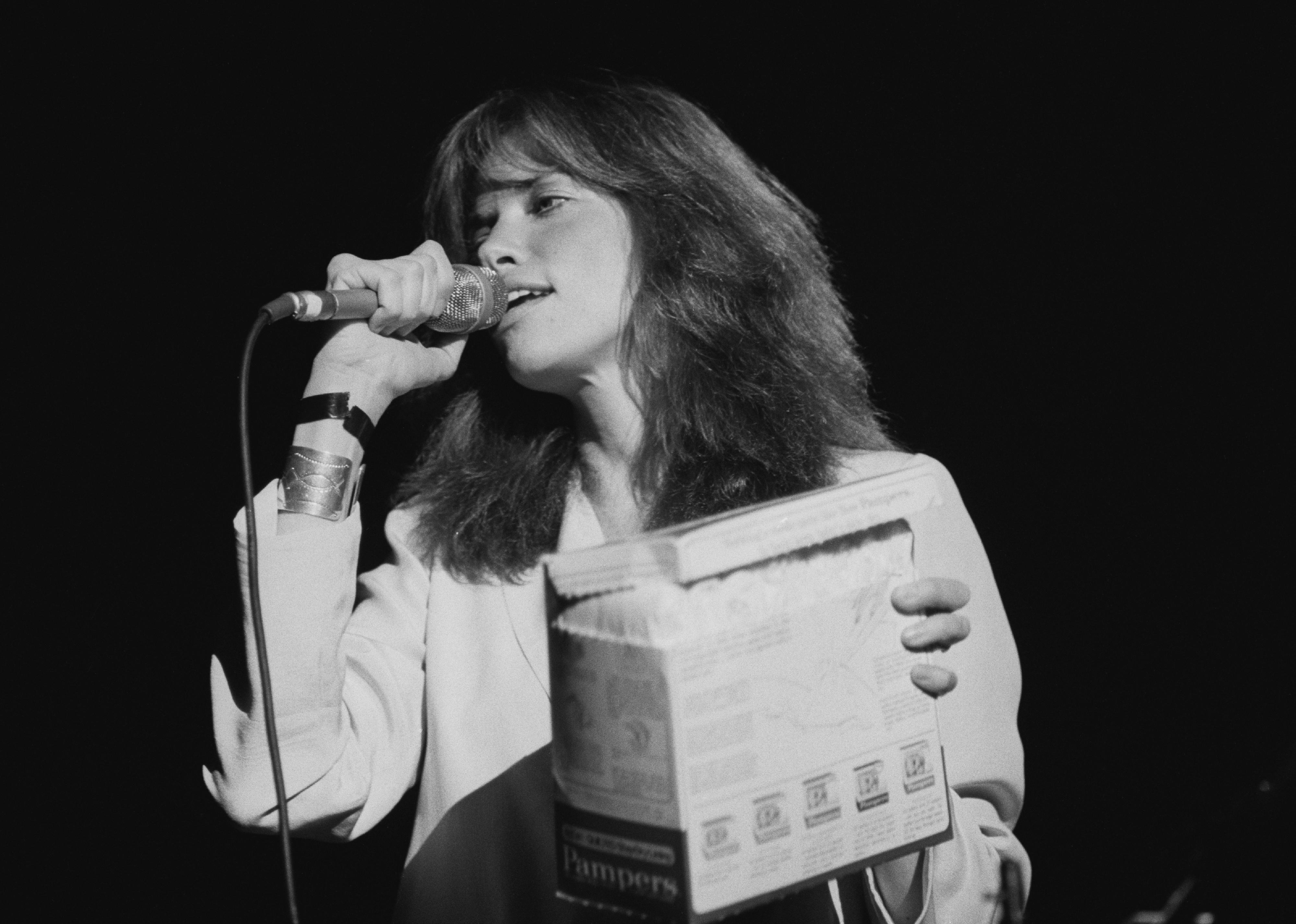 Carly Simon performing onstage holding a box of pampers.