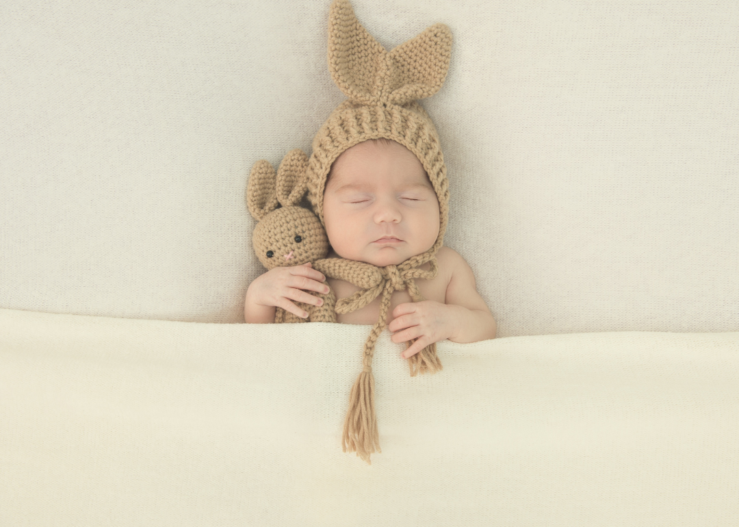 A sleeping baby in a brown hat with ears holding a bunny.