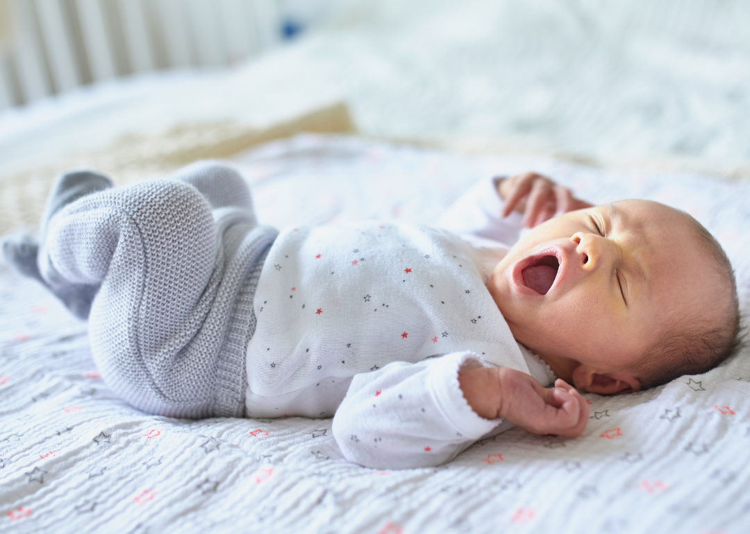 A baby lying in a crib wearing gray and white with stars and yawning.