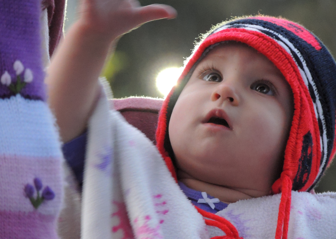 A baby in a red and blue hat and colorful blanket reaching up.