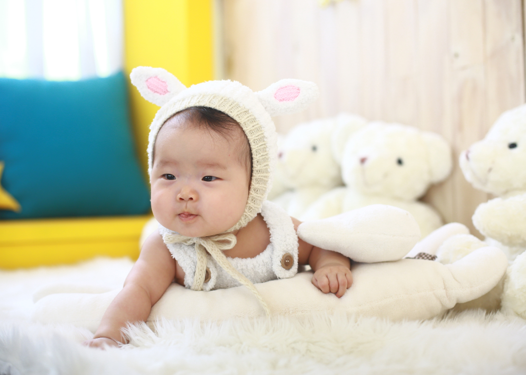 A baby girl in a white outfit and hat with ears surrounded by white teddy bears.
