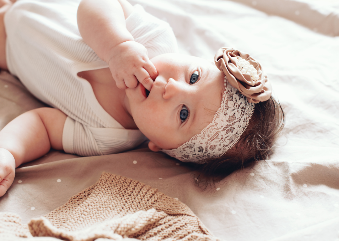 A baby girl with her hand in her mouth wearing a lace headband.