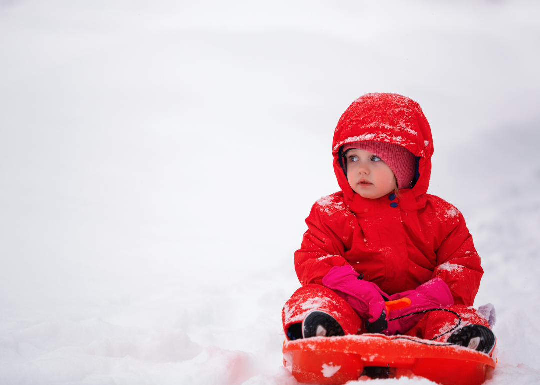 A baby girl in a pink hat and gloves and a red snow suit on a sled.