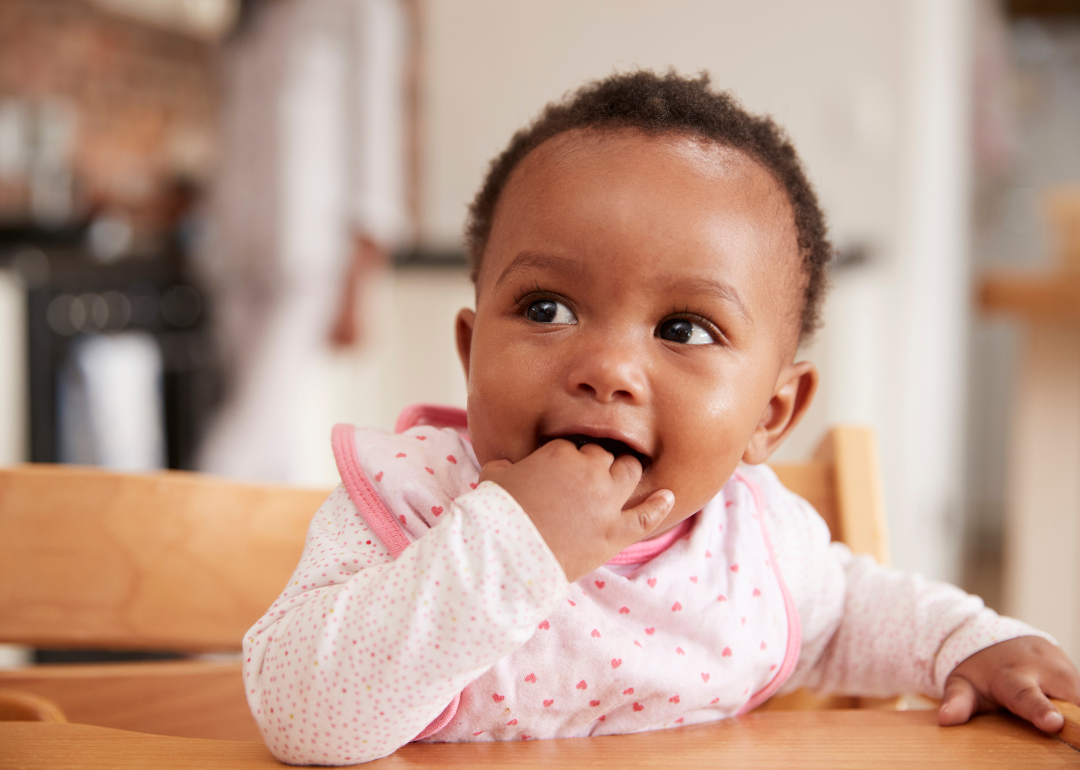 A baby girl in a high chair with her hand in her mouth.