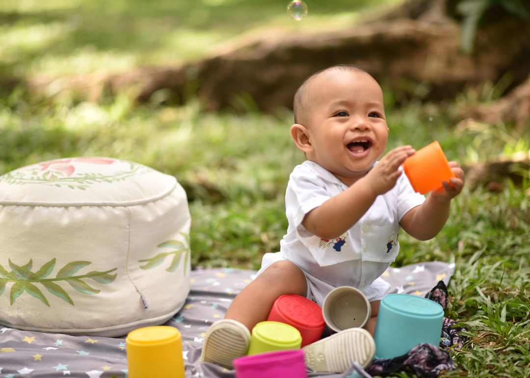 A smiling baby boy on a blanket outside playing with colorful cups.
