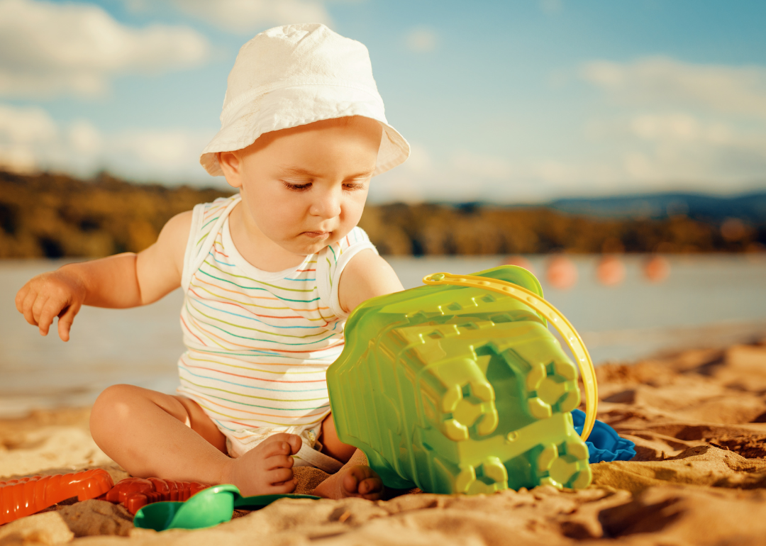 A baby girl in a striped suit and bucket hat playing in the sand.