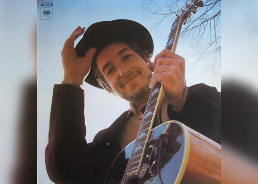 Bob Dylan looking down and smiling with a hat on holding a guitar.