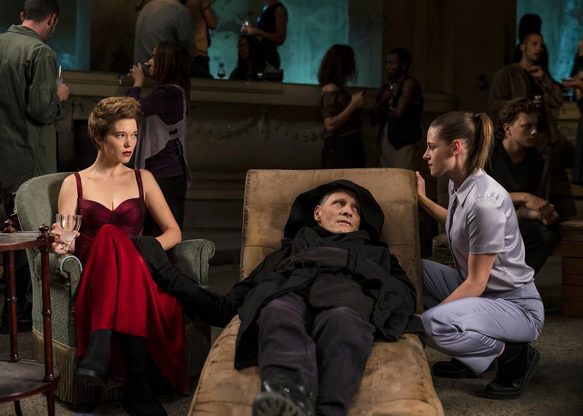 Viggo Mortensen lying on a lounge chair in a black straightjacket with Kristen Stewart and Léa Seydoux dressed up next to him