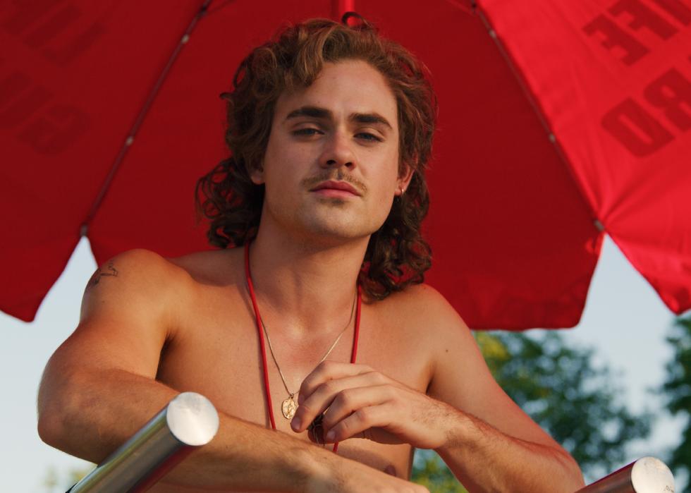 Dacre Montgomery in front of a big red umbrella in a lifeguard stand.