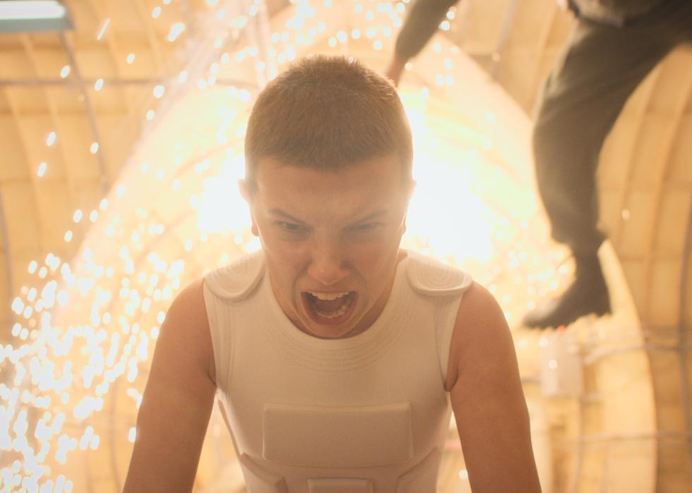 Millie Bobby Brown screaming in a tunnel with explosive light in the background and a person flying through the air.