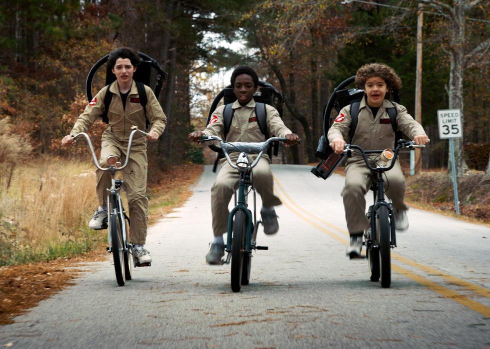 Caleb McLaughlin, Finn Wolfhard, and Gaten Matarazzo riding bikes in ghostbuster costumes.