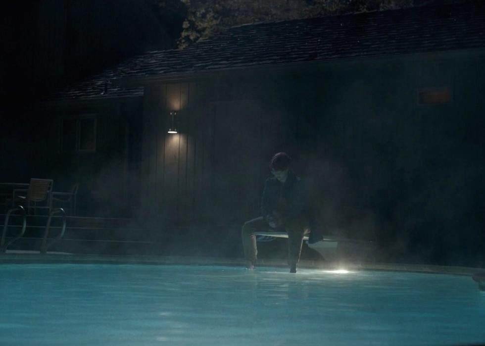 A girl sits on a diving board at night looking into a pool.