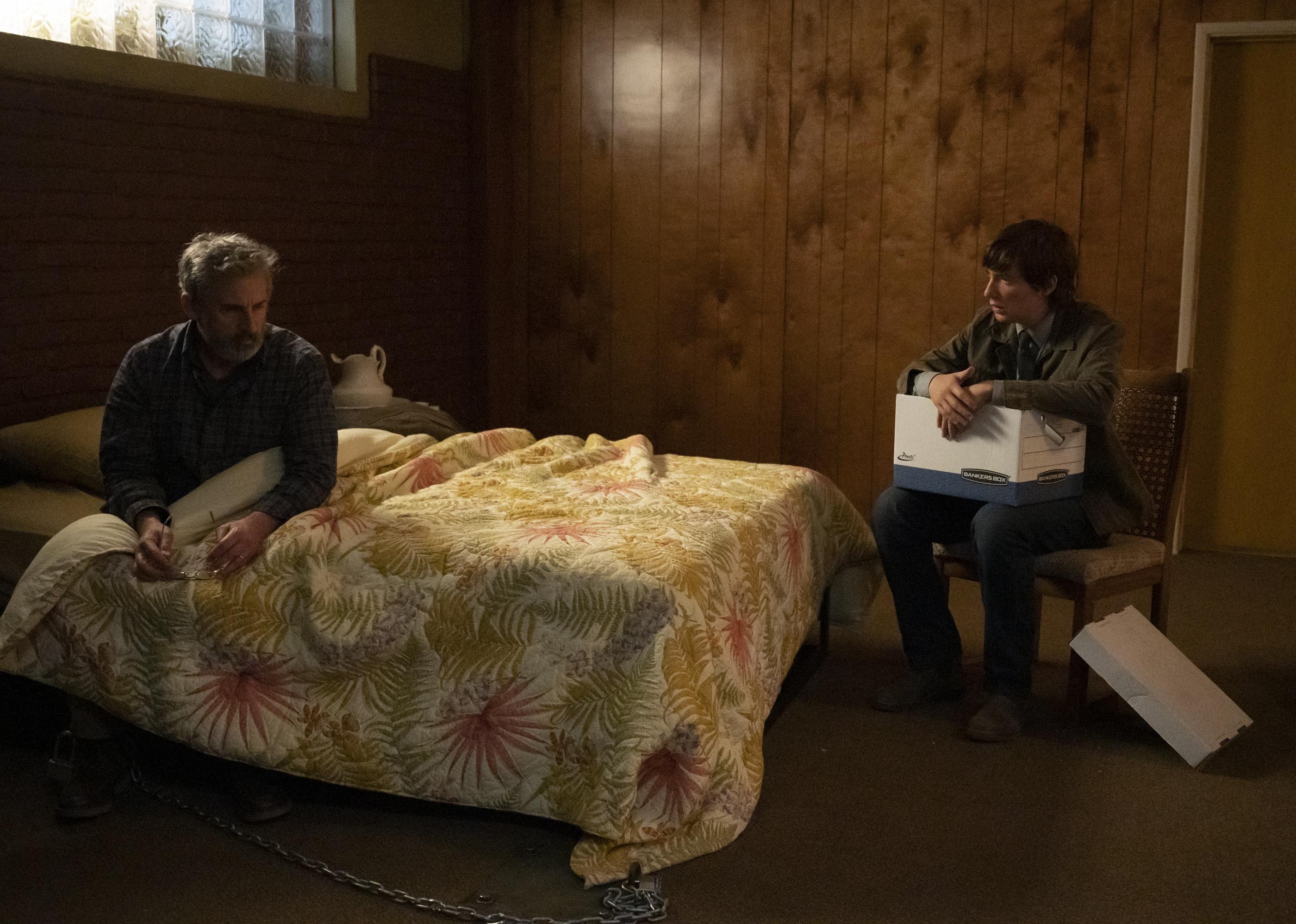 Steve Carell sitting chained to a bed as Domhnall Gleeson sits next to the bed in a chair holding a file box.