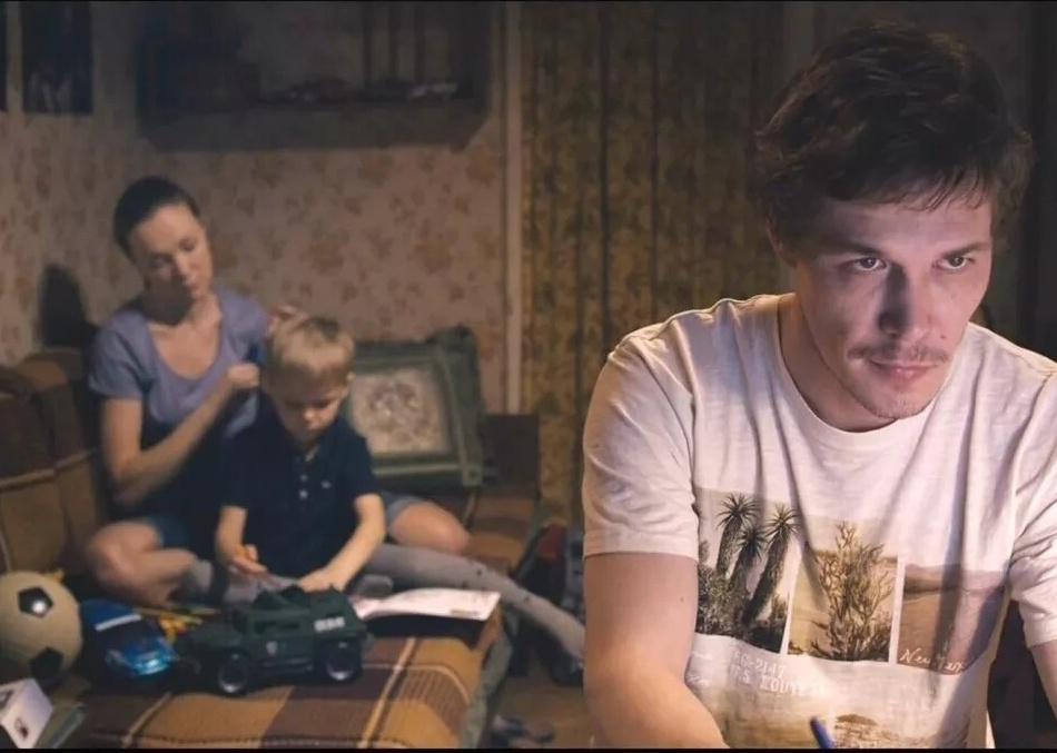 A man in a white t-shirt looks at a computer screen with a woman and child on a couch in the background.