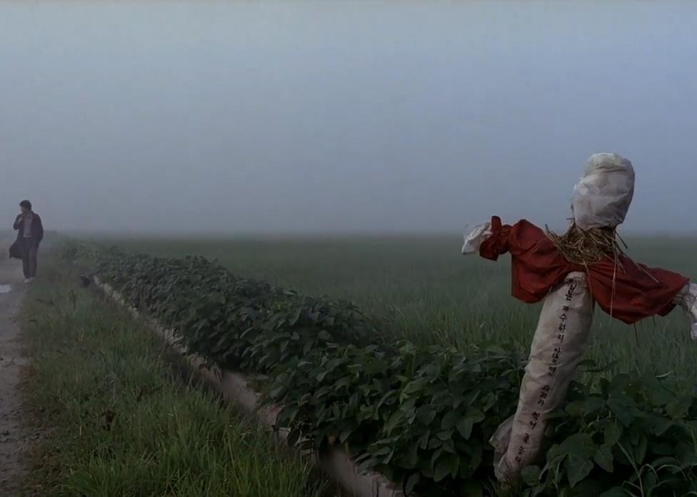 A man walking down a rural road in the fog with a scarecrow in the foreground.