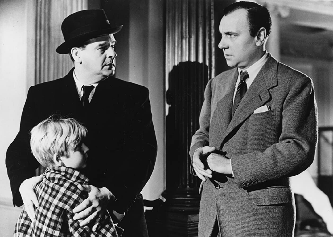 Two men in black and white stare at each other, one clutching a young boy at his feet.