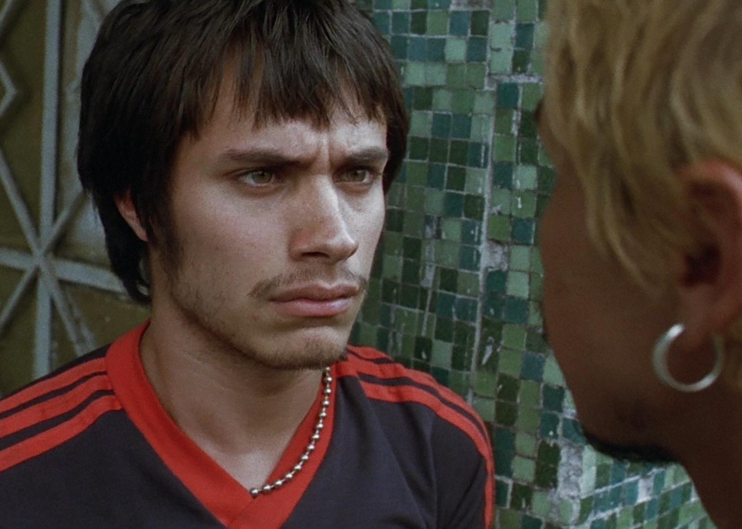Gael García Bernal in a black and red adidas shirt staring seriously at a man with blonde hair and an earring who is in his face.