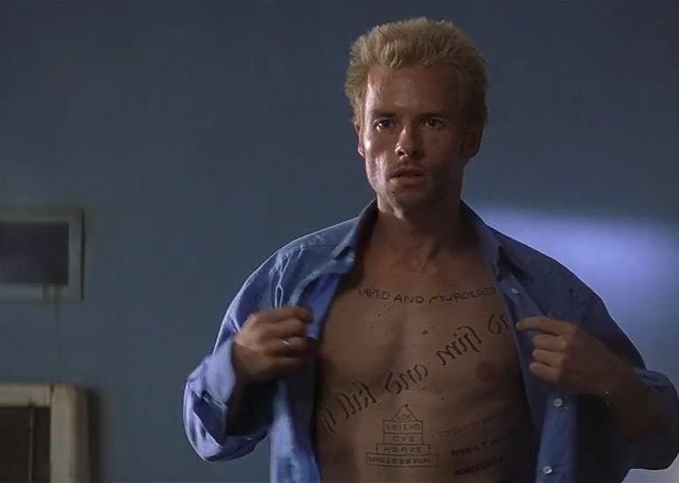 Guy Pearce with spikey blonde hair holding open his blue button down shirt reavealing writing all over his upper body.